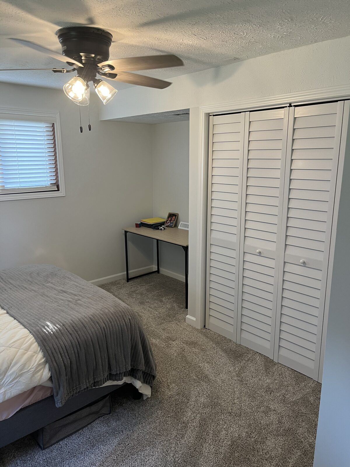 Finished basement bedroom with new carpet, white trim, louvered closet doors, ceiling fan, and a built-in desk nook