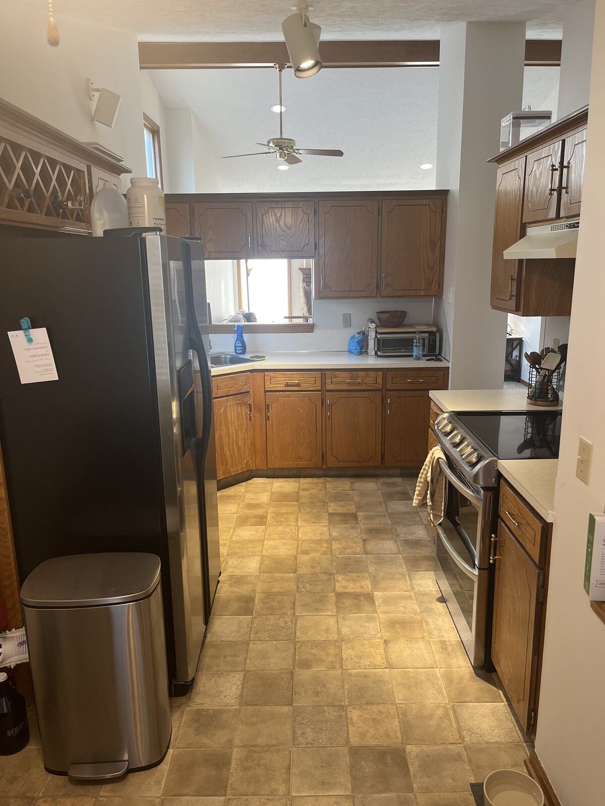 Original galley kitchen with dark oak cabinets, laminate counters, and dated tile-pattern vinyl floor looking toward the dining area