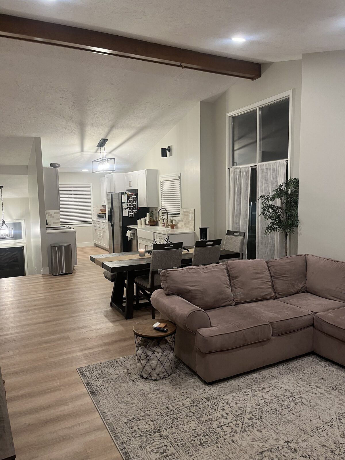 Open-concept living and dining space with wood-look flooring, exposed stained beam, lantern chandelier, and sightline into the renovated kitchen